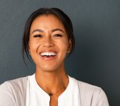 Smiling woman with dark hair against a dark gray background, wearing a white blouse and light cardigan.