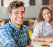 A man in a plaid shirt smiles while seated at a table in a cafe. A woman in a polka dot shirt is blurred in the background.