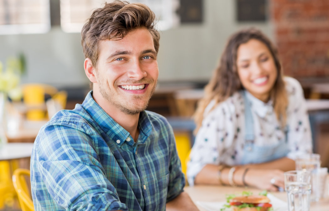 A man in a plaid shirt smiles while seated at a table in a cafe. A woman in a polka dot shirt is blurred in the background.