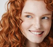 Person with wavy red hair smiling and looking to the side against a plain background.