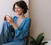 Woman in a blue blouse sitting against a wall, smiling at her phone. A potted plant is visible beside her.